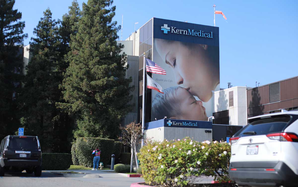 A shot outside Kern Medical showing the hospital building with a mural of a mother kissing a baby on the side. A flag pole with the US flag and the California flag flying.